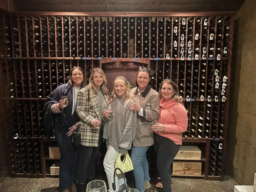 A tour group raising wine glasses in front of cellar bottle racks