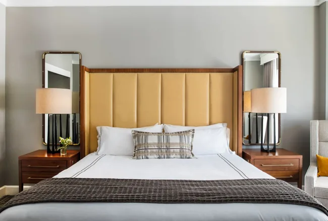 Deluxe King room overview with king bed, amber headboard, tufted bench, walnut nightstands, floor lamp, and large windows with drapes