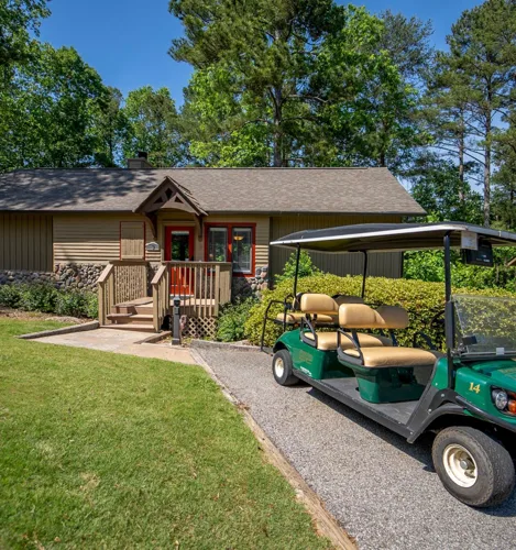 A small house with a golf cart parked in front, surrounded by greenery and tall trees under a sunny blue sky.